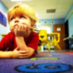 Photo description: Young toddler laying down on a carpet in a daycare.